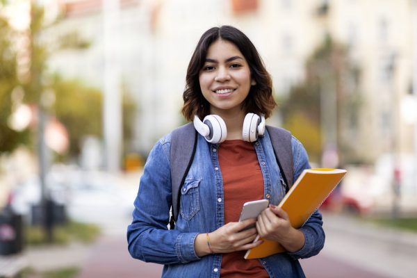 Modern Student. Portrait Of Smiling Young Arab Female With Workbooks And Smartphone In Hands Posing Outdoors, Beautiful Middle Eastern Woman With Backpack And Wireless Headphones Standing Outside